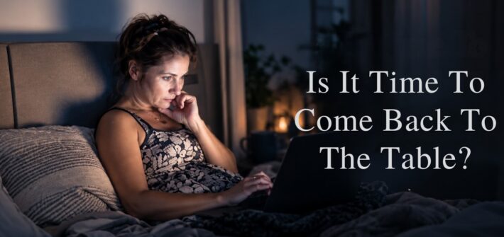 a woman sitting up in bed in a darkened bedroom, propped up on pillows and leaning against a headboard, Her face is illuminated only by a laptop computer screen.