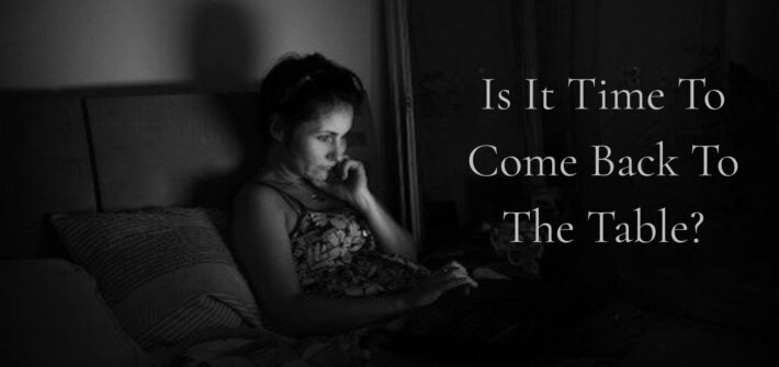A woman sitting in the darkness, staring at a phone screen. Text overlay Is it Time To Come Back To The Table