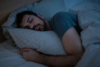 A young man, physically fit, peacefully sleeping on his stomach, hugging a pillow, in a darkened bedroom.