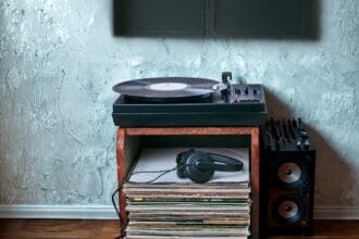 A record player on a stand; A pair of headphones sits on top of a stack of records on the shelf below. Background: A painted textured blue wall and a wooden floor.